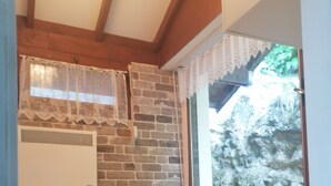 A bedroom with a brick wall, a window with lace curtains, a white bedside table with a lamp, and a bed with white bedding.