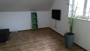 Een kamer met een houten vloer, een zwarte stoel, een flatscreen televisie aan de muur en een grote potplant.
