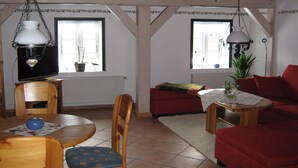 A cozy living room with a wooden dining table, red sofa, and a red armchair. There is a television on a stand and a potted plant on the windowsill. - North Sea Near Apartment under thatch (Ockholm)