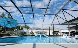 Zona de piscina con cielo despejado, una sombrilla azul y un edificio blanco al fondo.