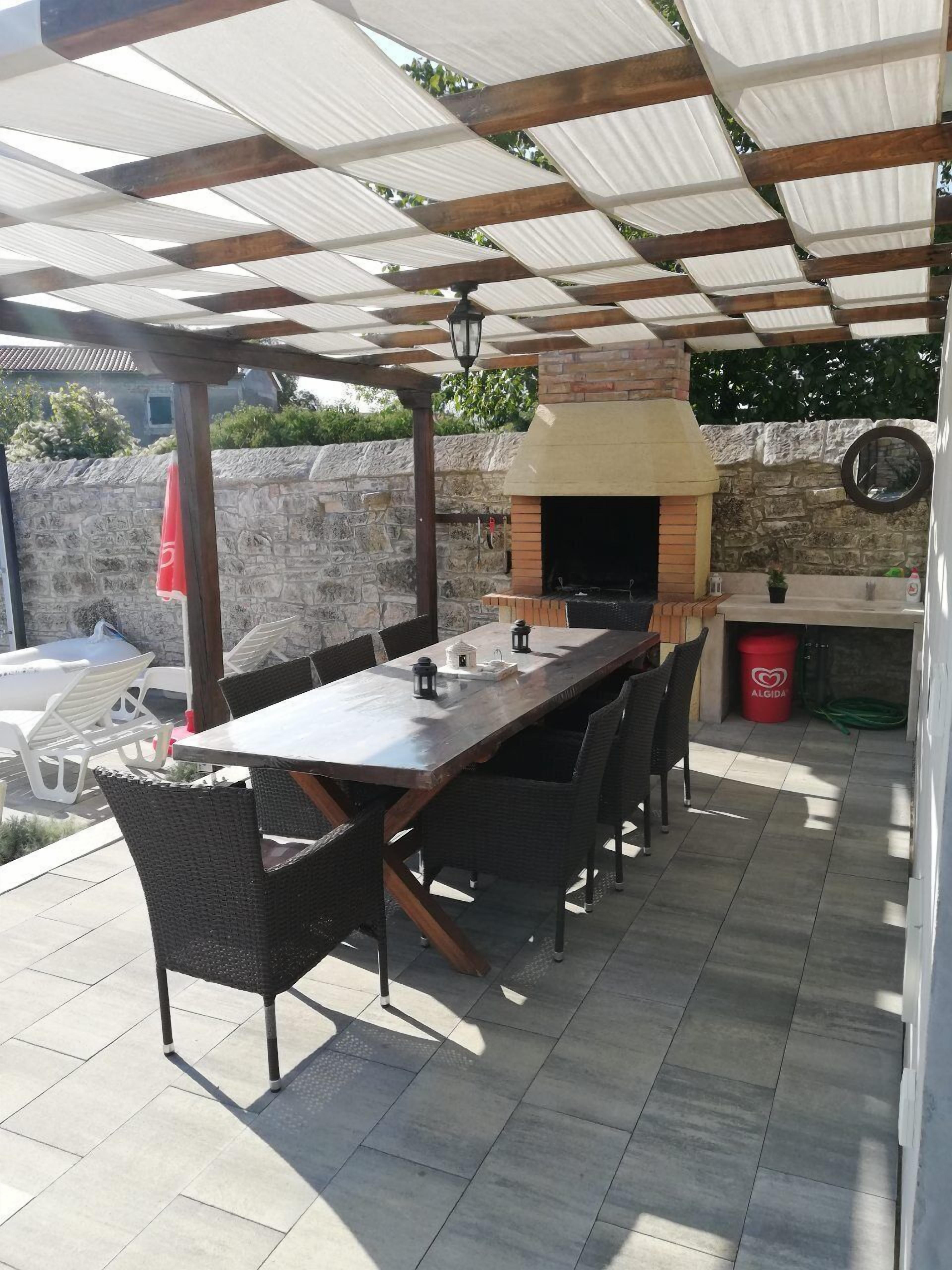 An outdoor patio with a long wooden table and wicker chairs, a stone wall, and a brick oven.
