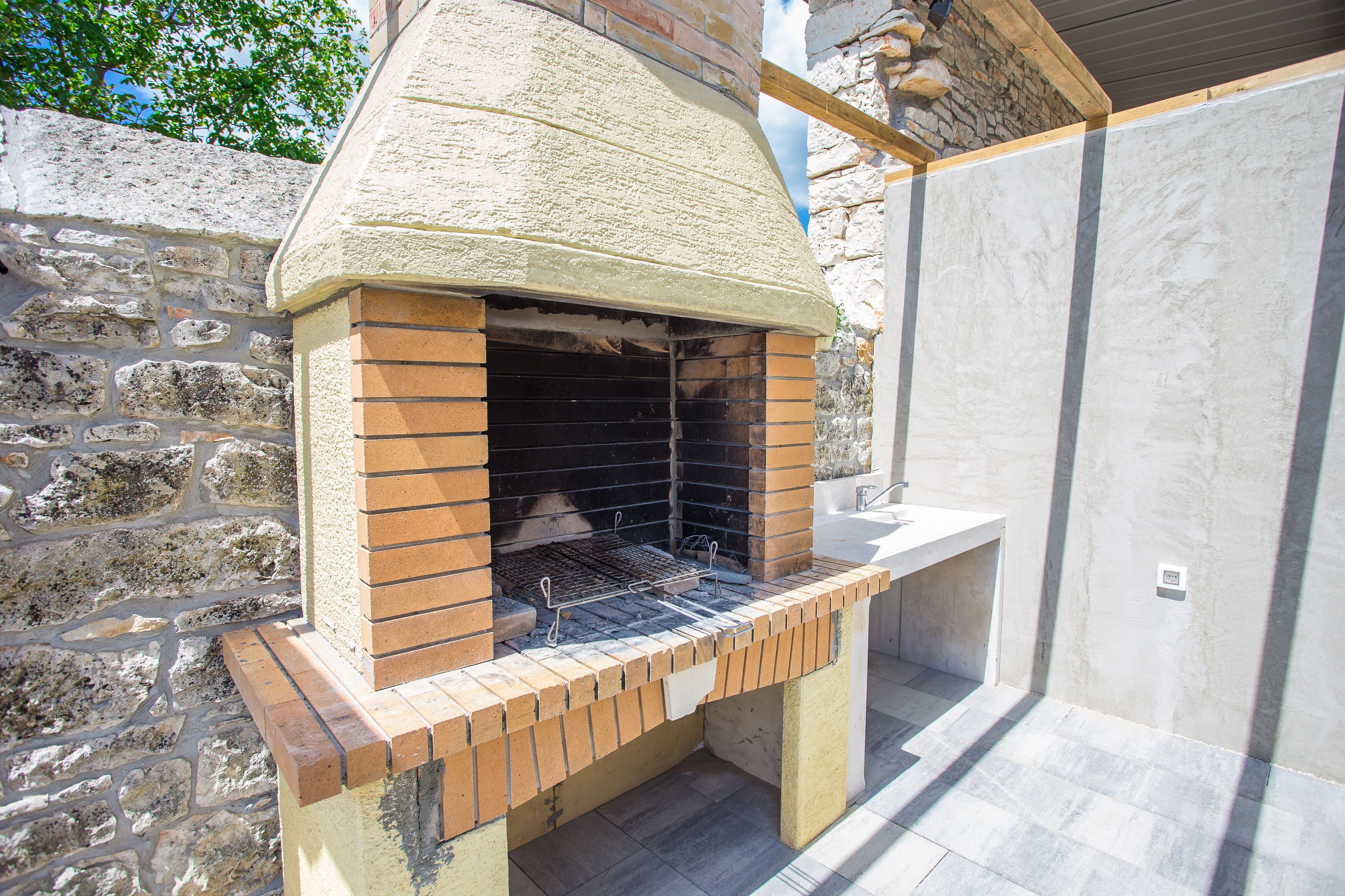 Un horno de ladrillo al aire libre con techo inclinado, ubicado junto a una pared de piedra y suelo de baldosas.
