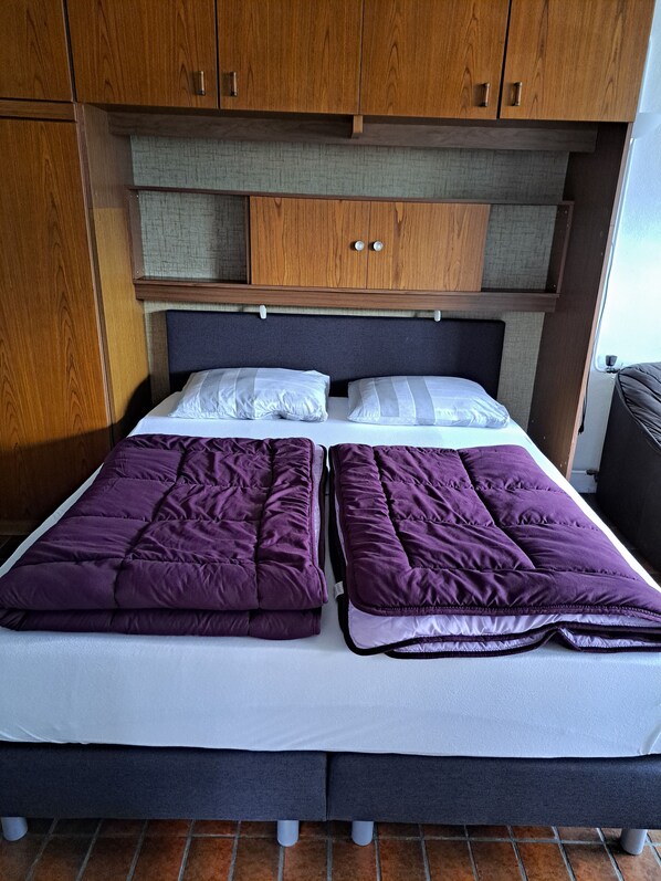 Una cama doble con dos edredones morados y almohadas blancas, armarios de madera arriba y piso de baldosas.
