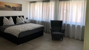 A bedroom with a large bed, a chair, a nightstand, a painting on the wall, and sheer curtains on the windows. - S-AN Daily Room Stuttgart Center (Claudio2) (Stuttgart Ost)
