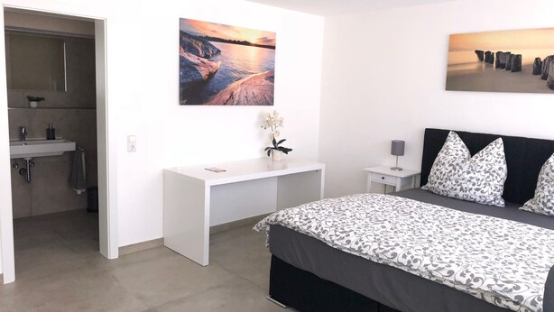 A bedroom with a bed, a desk, a chair, a nightstand, a lamp, and a painting on the wall. - S-AN Daily Room Stuttgart Center (Claudio2) (Stuttgart Ost)