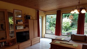 Un salon chaleureux avec un meuble TV en bois, un canapé et une table à manger. Une grande fenêtre, ornée de rideaux, offre une vue sur la verdure environnante.