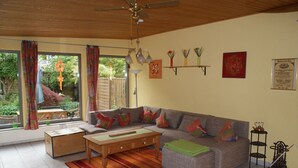 Un salon comprenant un canapé, une table basse et un petit meuble en bois. On y trouve une fenêtre avec des rideaux, un ventilateur de plafond et une étagère fixée au mur, ornée d’objets décoratifs.