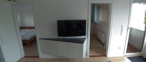 Une pièce compacte comprenant un lit, un téléviseur posé sur un meuble, une petite kitchenette et une porte-fenêtre coulissante donnant sur un espace extérieur.