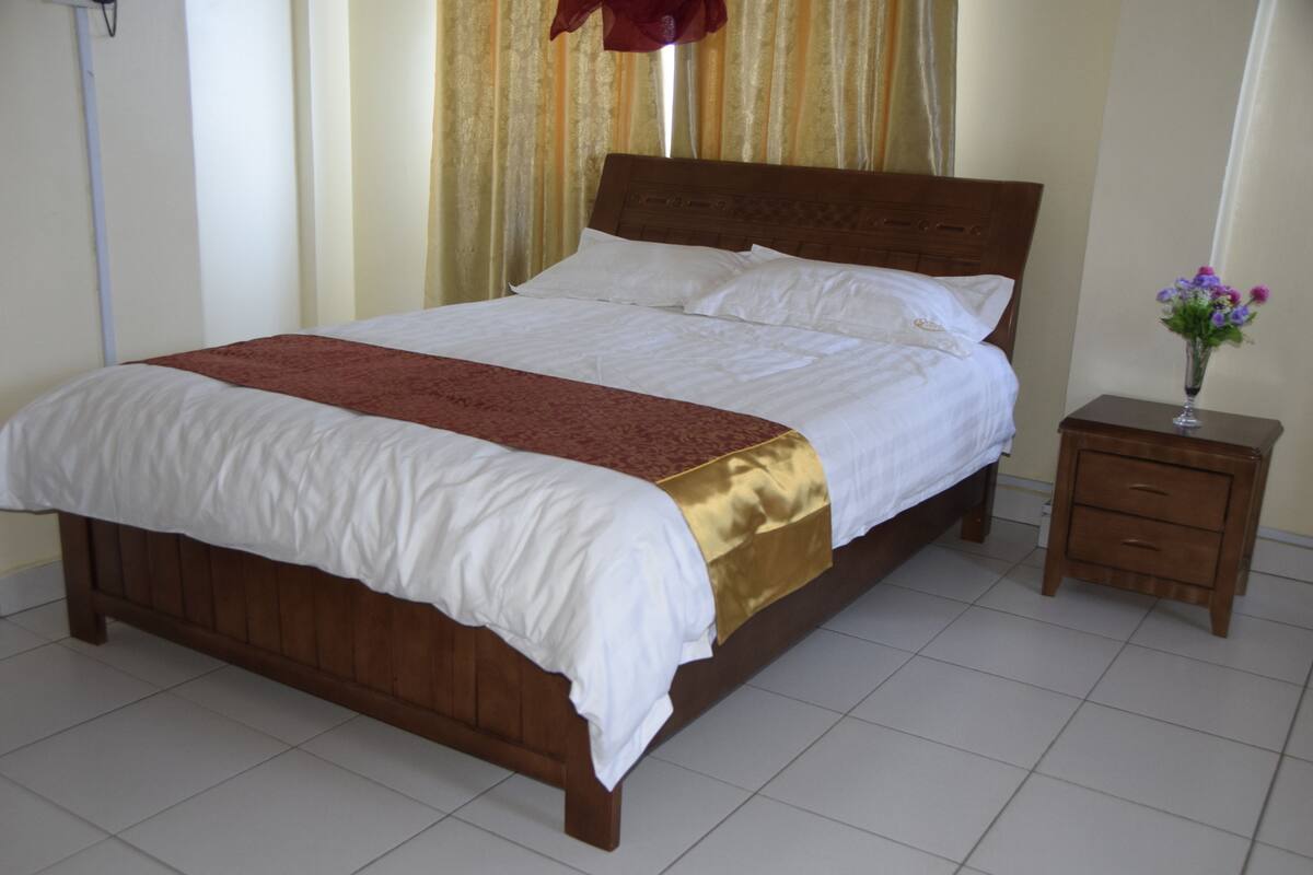 Een keurig opgemaakt bed met een houten hoofdeinde, een nachtkastje met lades en een vaas met bloemen.