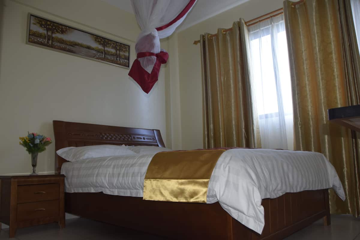 Een net opgemaakt bed met gouden accenten, een nachtkastje met een bloevas en een schilderij aan de muur.