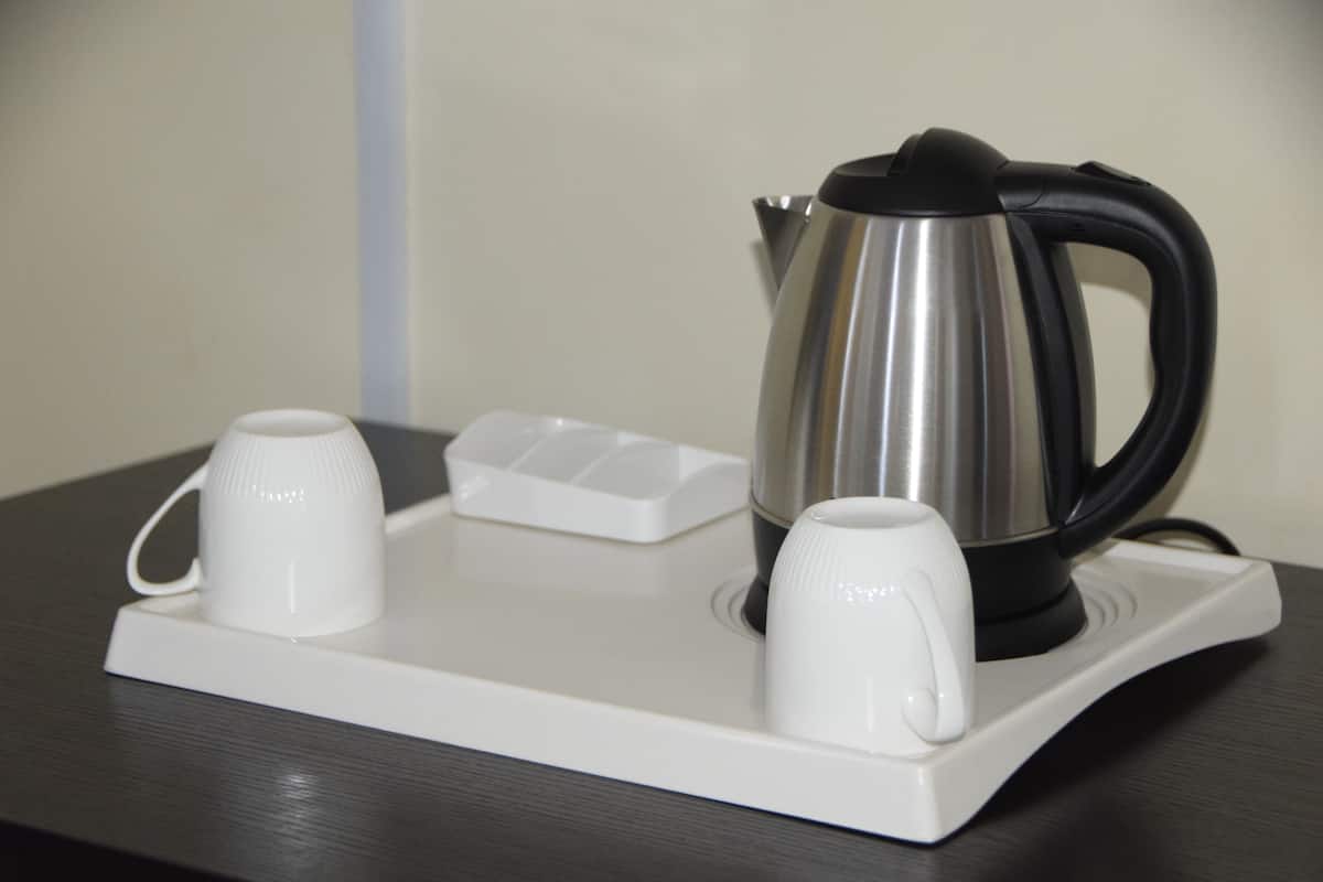 Een roestvrijstalen waterkoker met een zwarte hendel op een witte schaal met twee witte bekers.