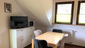 Une petite pièce bien éclairée, avec une table à manger, des chaises, un téléviseur et un four à micro-ondes. Il y a une fenêtre avec des stores et un radiateur en dessous.