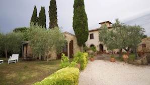 Eine Villa mit Kopfsteinpflasterauffahrt, Sitzgelegenheiten im Freien und einem Garten mit Kübelpflanzen und hohen Bäumen.