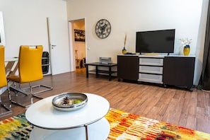 A modern living room with a flat-screen TV, a clock, and a dining area with yellow chairs.