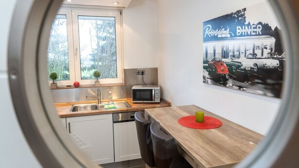 Une cuisine moderne équipée d’un four à micro-ondes, d’un évier et d’un plan de travail en bois. On aperçoit une table à manger avec des chaises, et un tableau représentant des voitures anciennes est accroché au mur.