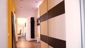 Un couloir avec un plancher en bois, un mur jaune et un mur à rayures noires et blanches. On y voit une porte menant à une autre pièce, un vase blanc avec des fleurs jaunes et une chaise au fond.
