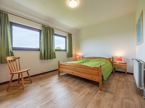 A bedroom with a wooden bed, a chair, two windows with curtains, a bedside table with a lamp, and a radiator.