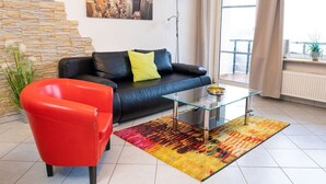 Un salon avec un canapé en cuir noir, un fauteuil rouge, une table basse en verre et un tapis aux couleurs vives.