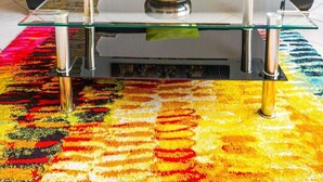 Un salon moderne avec un canapé en cuir noir, une table basse en verre et un tapis aux motifs éclatants. Un tableau représentant un paysage urbain est accroché au mur, et un escalier est visible sur la gauche.