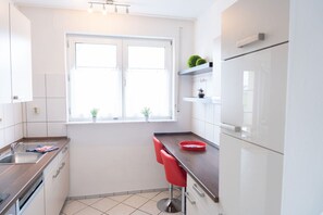 Una cucina moderna con mobili bianchi, lavello in acciaio inossidabile, una finestra e due piante in vaso, e uno sgabello rosso.