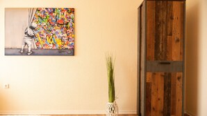 Une pièce avec un meuble en bois, un vase blanc contenant des plantes vertes et, sur le mur, un tableau coloré dans le style du graffiti.