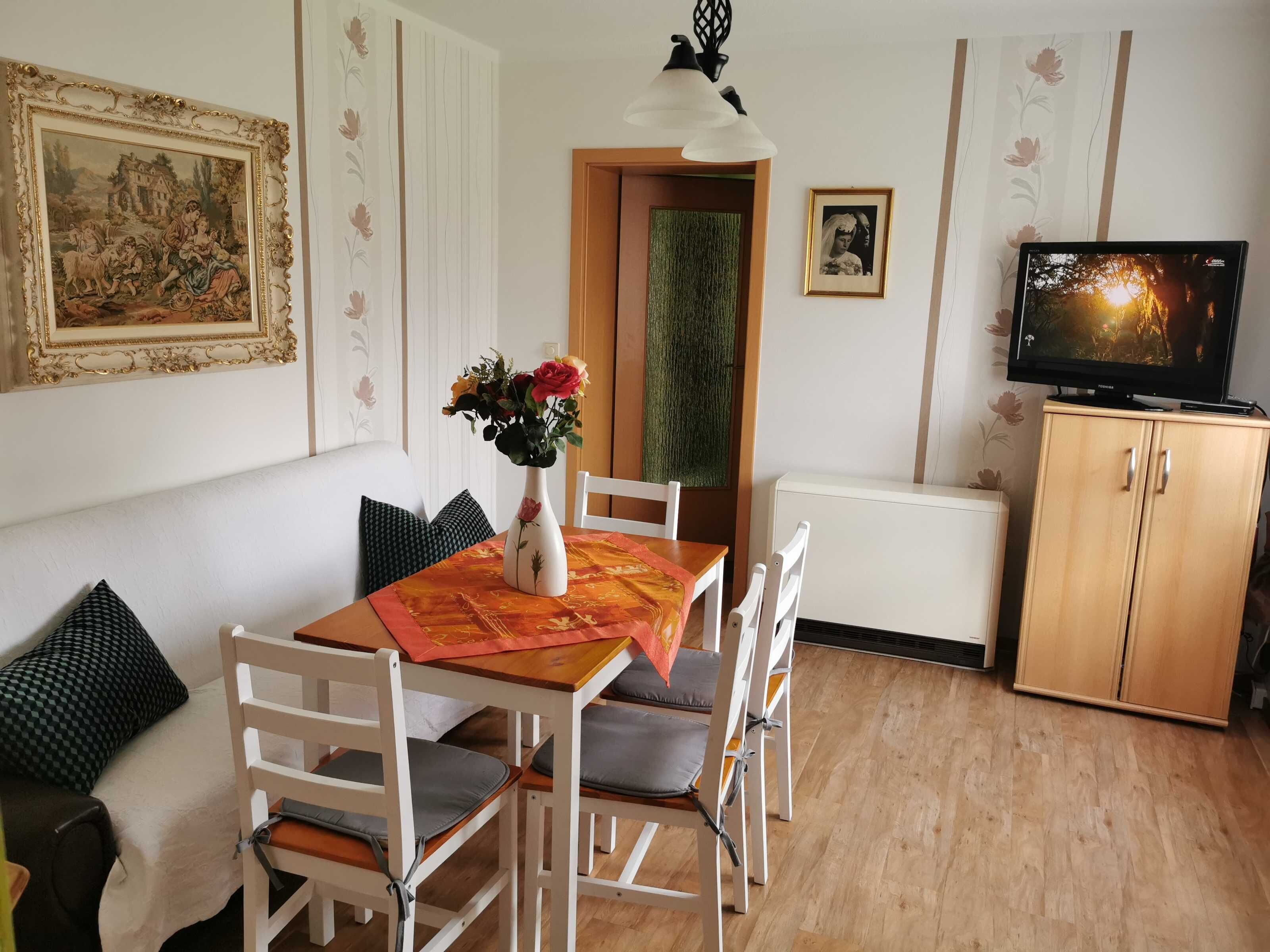 En matplats med ett bord, stolar, en vas med blommor, en tavla, en tv och ett skåp.