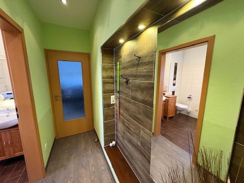 Un couloir doté d’un plancher en bois, d’une porte en verre et d’un miroir qui reflète une salle de bain avec des toilettes et un lavabo.