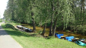 Un área de picnic a orillas del río, con mesas y sillas, una embarcación atracada y un sendero a lo largo de la orilla.