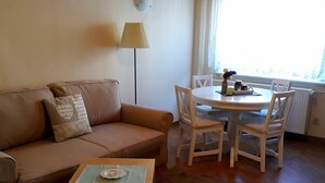 Un salón con un sofá color beige, una mesita de madera con una vela, sillas blancas, una lámpara y una ventana con cortinas.