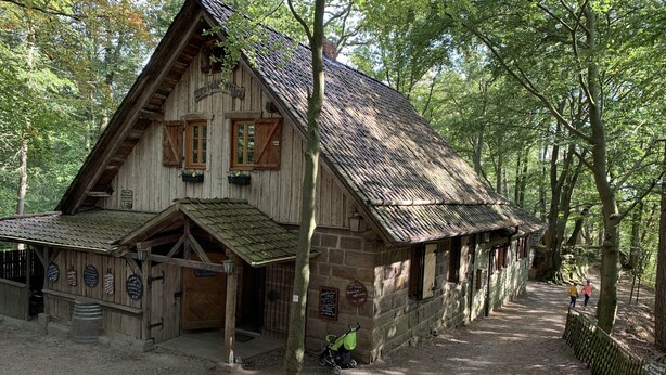 A rustic wooden building with a steeply pitched roof, a front porch, and a dirt path leading to it.