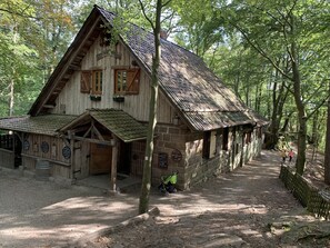 Een rustiek houten gebouw met een steil dak, een veranda en een onverharde pad er naartoe.