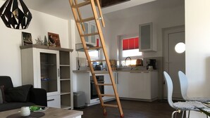 A modern living room with a wooden coffee table, a sofa, and a white cabinet. A wooden ladder is leaning against the wall, and there is a kitchen area with white cabinets and a red window blind.