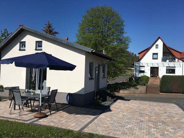 Een huis met een patio, voorzien van een tafel, stoelen, een blauwe parasol en een tuinslang.