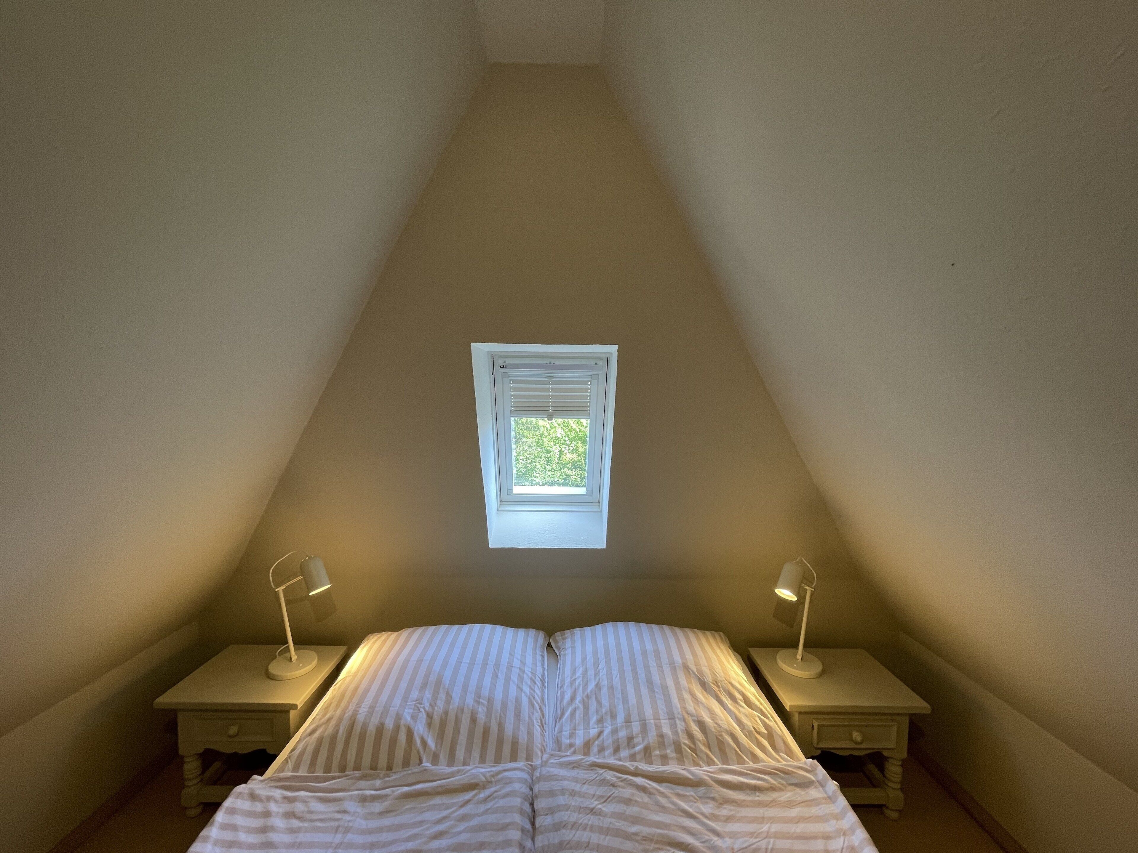 Et soveværelse med skråvægg, et vindue, to sengeborde med lamper, og en seng med stribet sengetøj.
