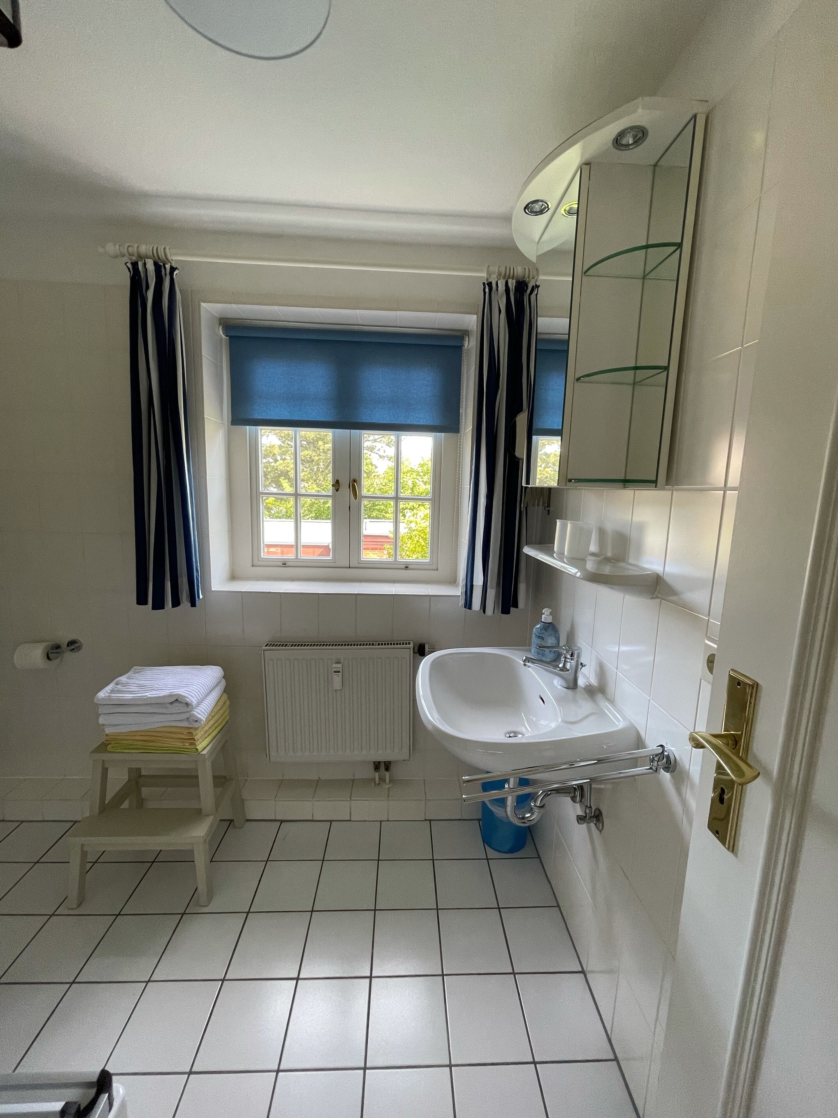 A bathroom with a white sink, a window with blue curtains, a radiator, and a small stool with towels.