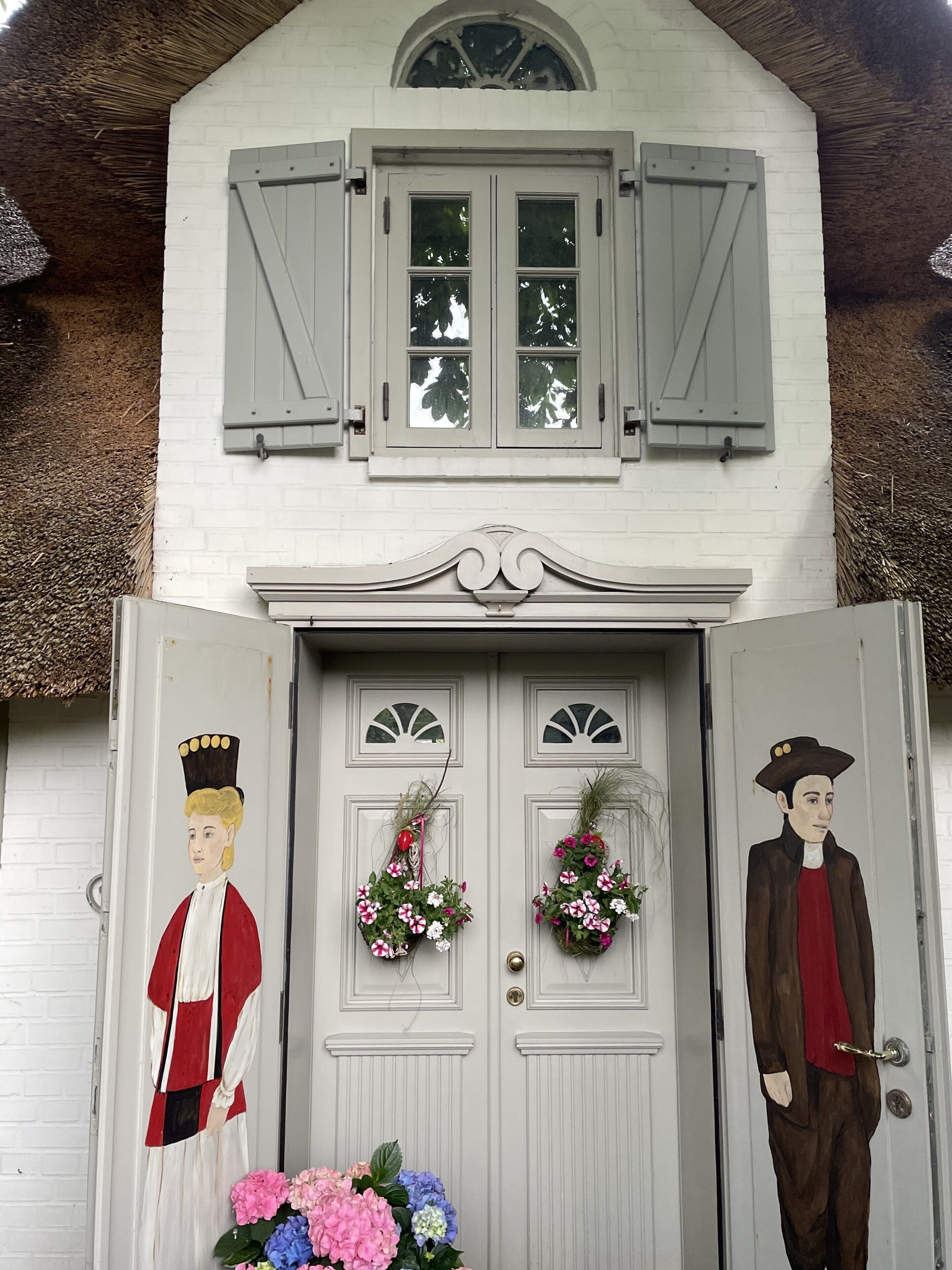 A white building with a thatched roof, a double door with two windows, and two door knockers with painted figures.