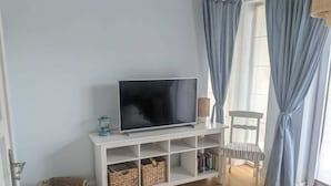 Un salón con televisor de pantalla plana sobre un mueble blanco, cestos de mimbre, un sillón de mimbre y una ventana con cortinas azules.