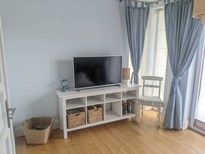 Ein Wohnzimmer mit einem Flachbildfernseher auf einem weißen TV-Schrank, Korbwannen, einem Korbstuhl und einem Fenster mit blauen Vorhängen.