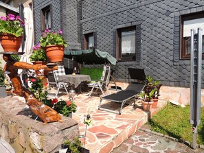 Un patio con una mesa, sillas y una silla de descanso. Hay plantas en macetas y una estructura de madera decorativa.