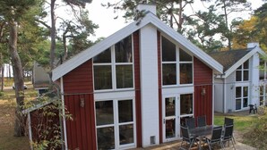 A small, single-story house with a red exterior and white trim, featuring large windows and a patio with outdoor furniture. - House in the dunes (Mecklenburg Vorpommern)