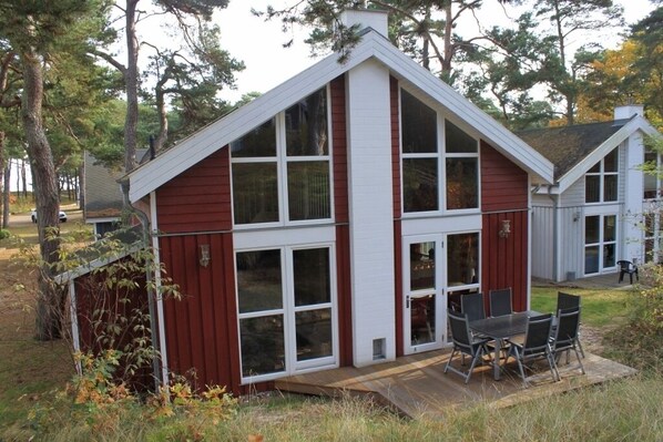 A small, single-story house with a red exterior and white trim, featuring large windows and a patio with outdoor furniture. - House in the dunes (Mecklenburg Vorpommern)