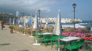 Ein Straßencafé mit Tischen, Stühlen und Sonnenschirmen, mit Blick auf das Meer und Gebäude im Hintergrund.