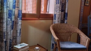 Ein Zimmer mit einem Korbstuhl, einem kleinen Holztisch mit Büchern und einem Fenster mit gemusterten Vorhängen.