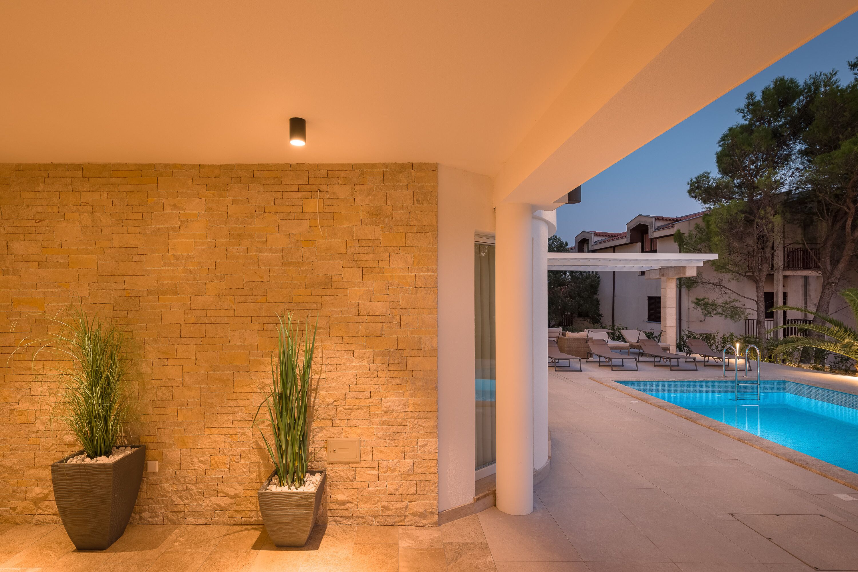 A pool area with a brick wall, two large planters with plants, and a view of a building in the background.