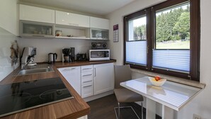 Een moderne keuken met een ingebouwde kookplaat, magnetron en koffieapparaat. Er is een eettafel met een schaal fruit en een raam met jaloezieën.