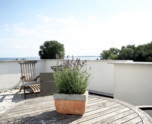 Eine Holzterrasse mit einem runden Tisch, Stühlen, einem Blumentopf und Blick auf das Meer.