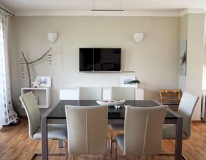 A modern dining area with a rectangular table, six chairs, a flat-screen TV mounted on the wall, and a small side table with a plant. - House at the sea (Wohlenberg)