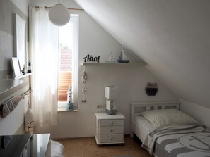 A bedroom with a sloped ceiling, a bed with a nautical-themed bedspread, a nightstand with a lamp, and a window with a view of a red-roofed building. - House at the sea (Wohlenberg)