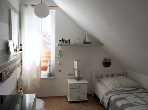 A bedroom with a sloped ceiling, a bed with a nautical-themed bedspread, a nightstand with a lamp, and a window with a view of a red-roofed building. - House at the sea (Wohlenberg)