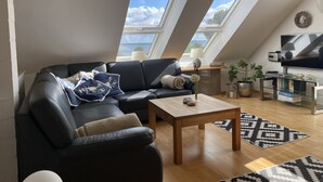 A modern loft apartment with a black leather sofa, a wooden coffee table, and a TV on a stand. There are two skylights, a lamp, and a bookshelf with books. - House at the sea (Wohlenberg)